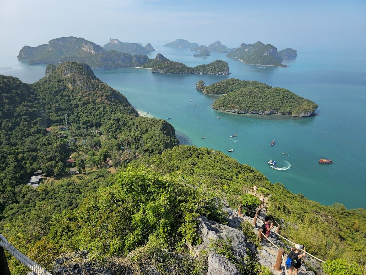 Panoramic view from a hilltop in Ang Thong National Marine Park over a cluster of forested limestone islands rising from turquoise Gulf of Thailand water, with hikers on a rocky ridge in the foreground