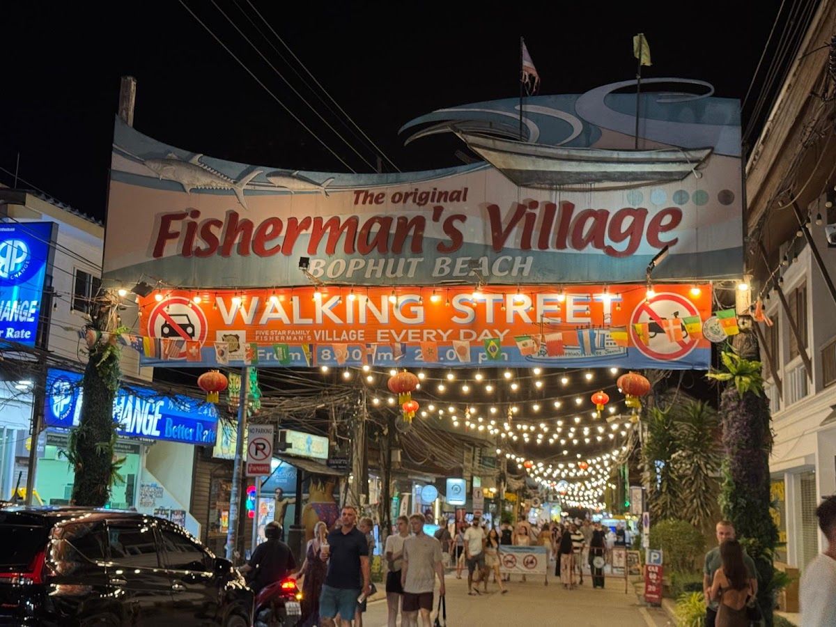 Fisherman's Village Walking Street entrance at Bophut Beach, Koh Samui — festive string lights stretching over Beach Road, red lanterns, and visitors browsing the Friday night market