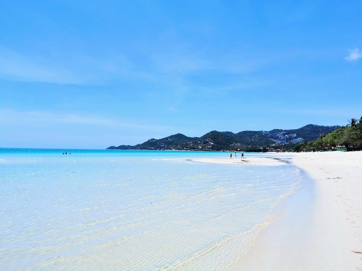 Chaweng Beach, Koh Samui — long white-sand beach stretching north with turquoise water, sun loungers and the beachfront hotel strip backed by green hills