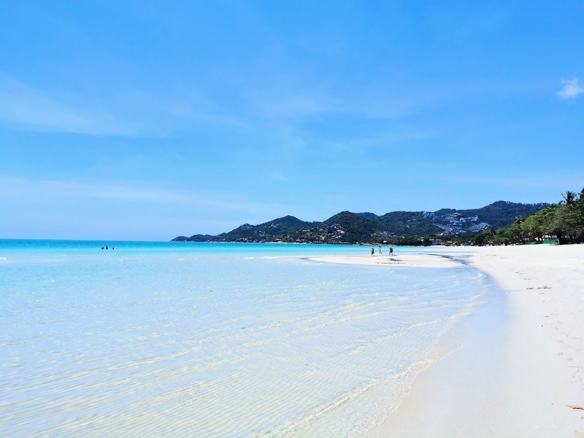 Chaweng Beach, Koh Samui on a clear day: wide stretch of white sand with bright turquoise water and green wooded hills in the background