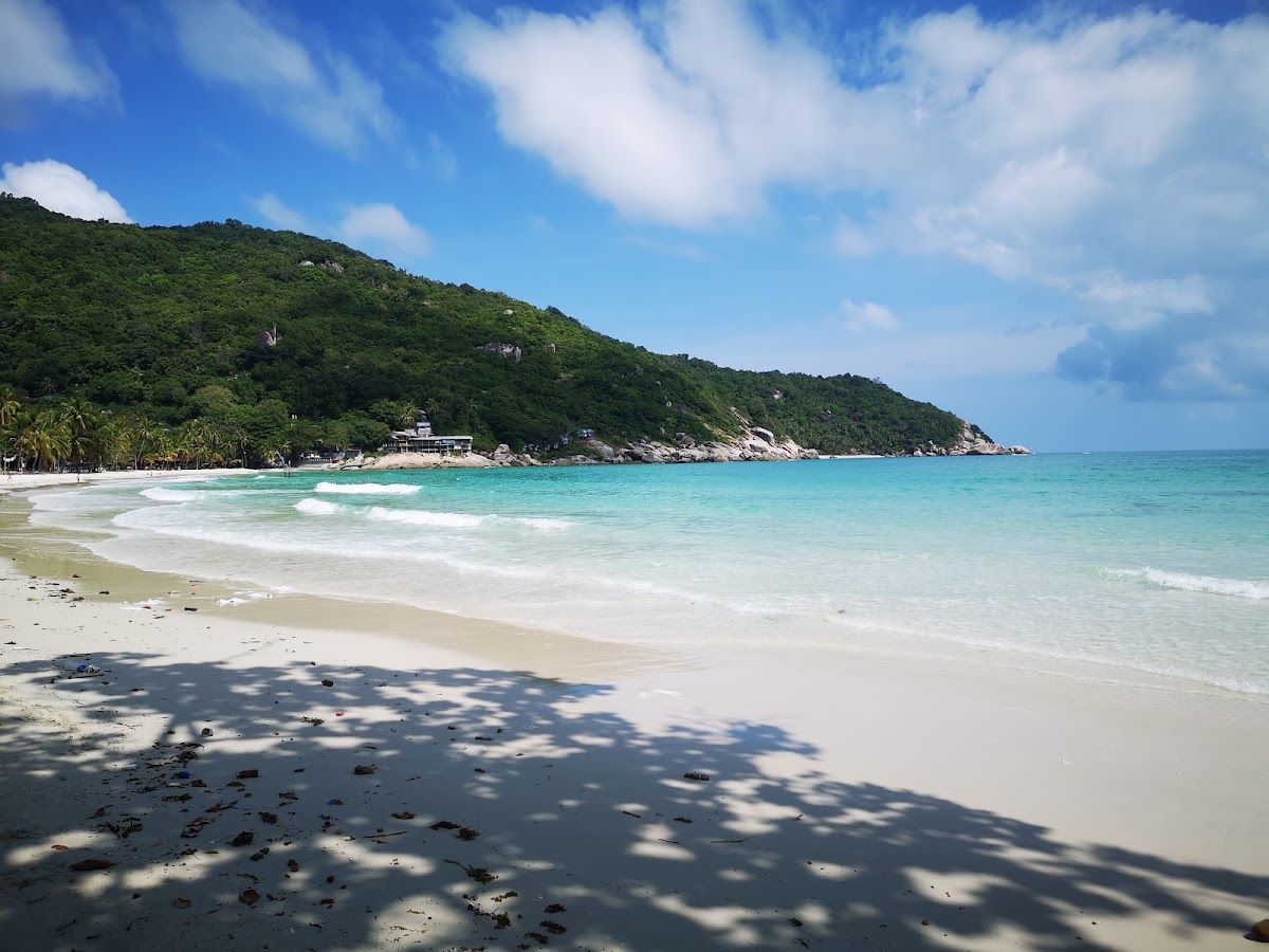 Haad Rin beach on Koh Phangan in daylight — white sand, turquoise Gulf water and jungle hills, the site of the Full Moon Party