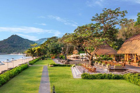 Vana Belle Koh Samui resort — manicured tropical gardens leading to a white sand beach with calm Gulf of Thailand water and forested hills beyond