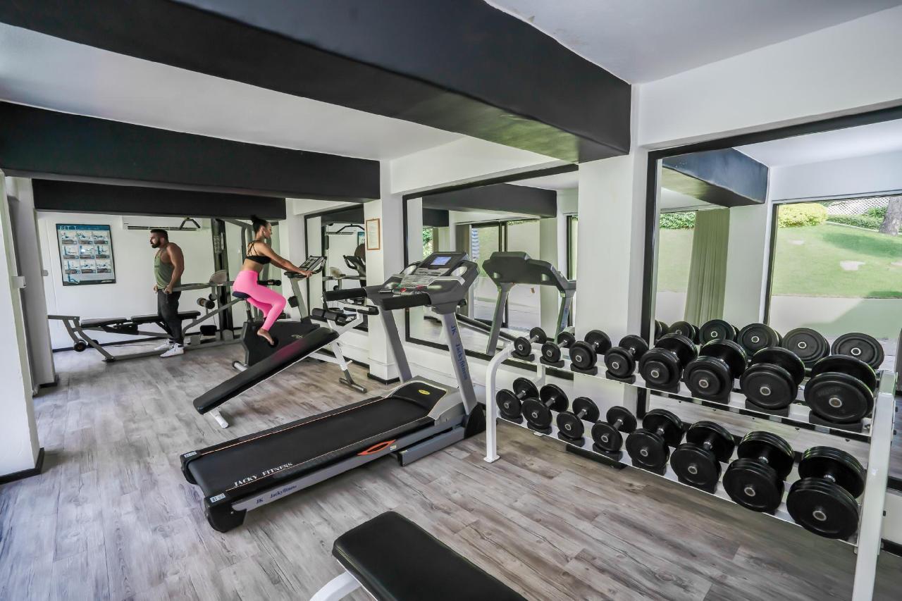 Gym at The Briza Beach Resort, with weight machines, dumbbells and a window toward the trees